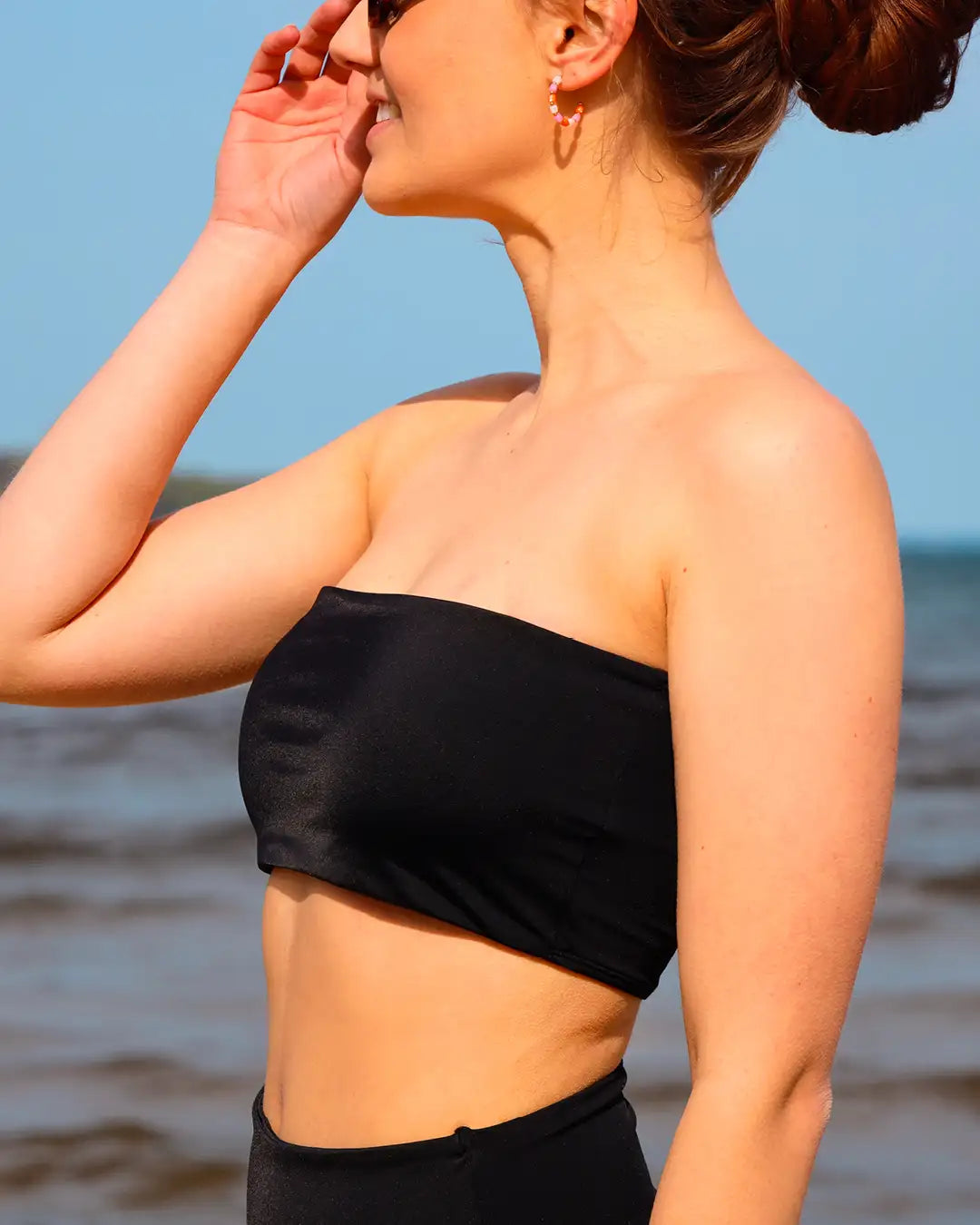 Eine Frau, am Meer, in einem schwarzen Bandeau Bikini richtet ihre Sonnenbrille.