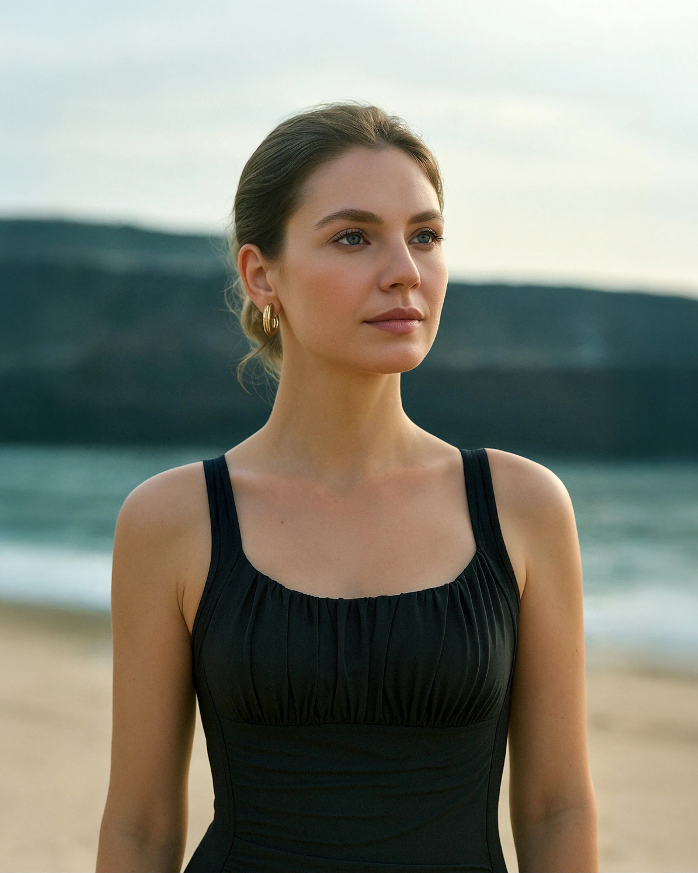 Eine junge Frau steht am Strand und schaut in die Ferne. Sie trägt den schwarzen Shape Badeanzug von MYLILY