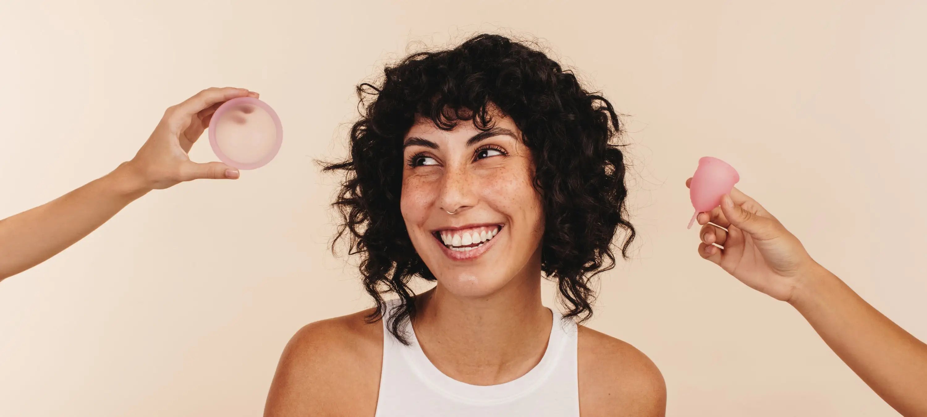 Man sieht eine lächelnde Frau mit locken und zwei Arme, die eine hält eine Menstruationsscheibe und die andere eine Menstruationstasse. Es handelt sich um eine Gegenüberstellung