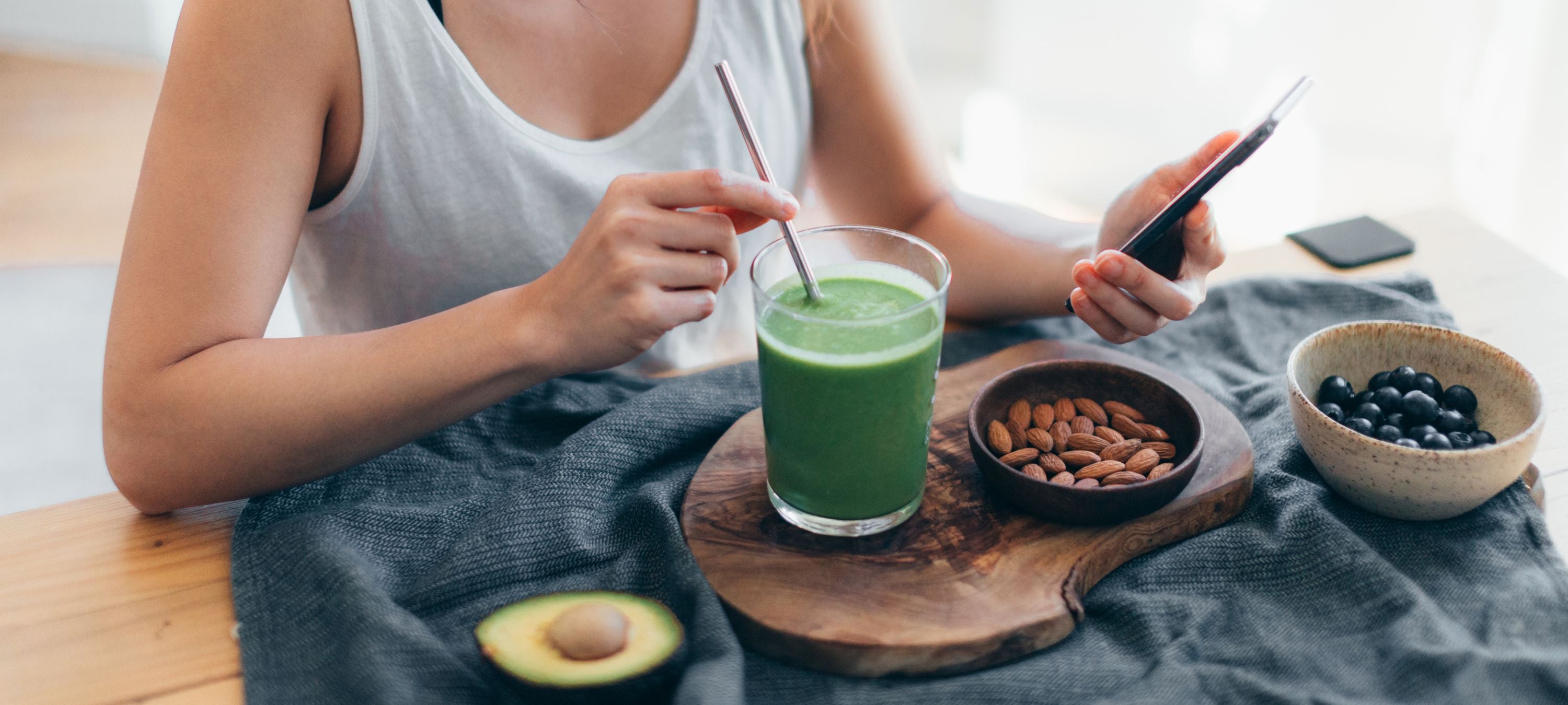 Frau trinkt grünen Smoothie und sitzt an Tisch mit veganen Lebensmitteln