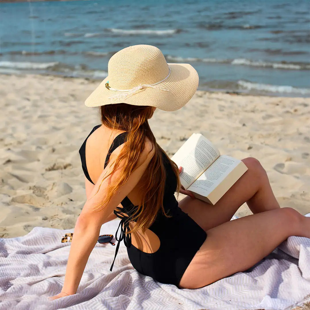 Junge Frau mit Sonnenhut und Perioden Badeanzug liest am Strand auf einem Handtuch.