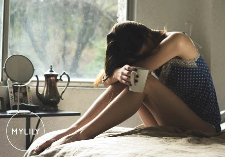 Eine Frau sitzt zusammengekrümmt auf der Ecke ihres Bettes und hält eine Tasse in der rechten Hand.