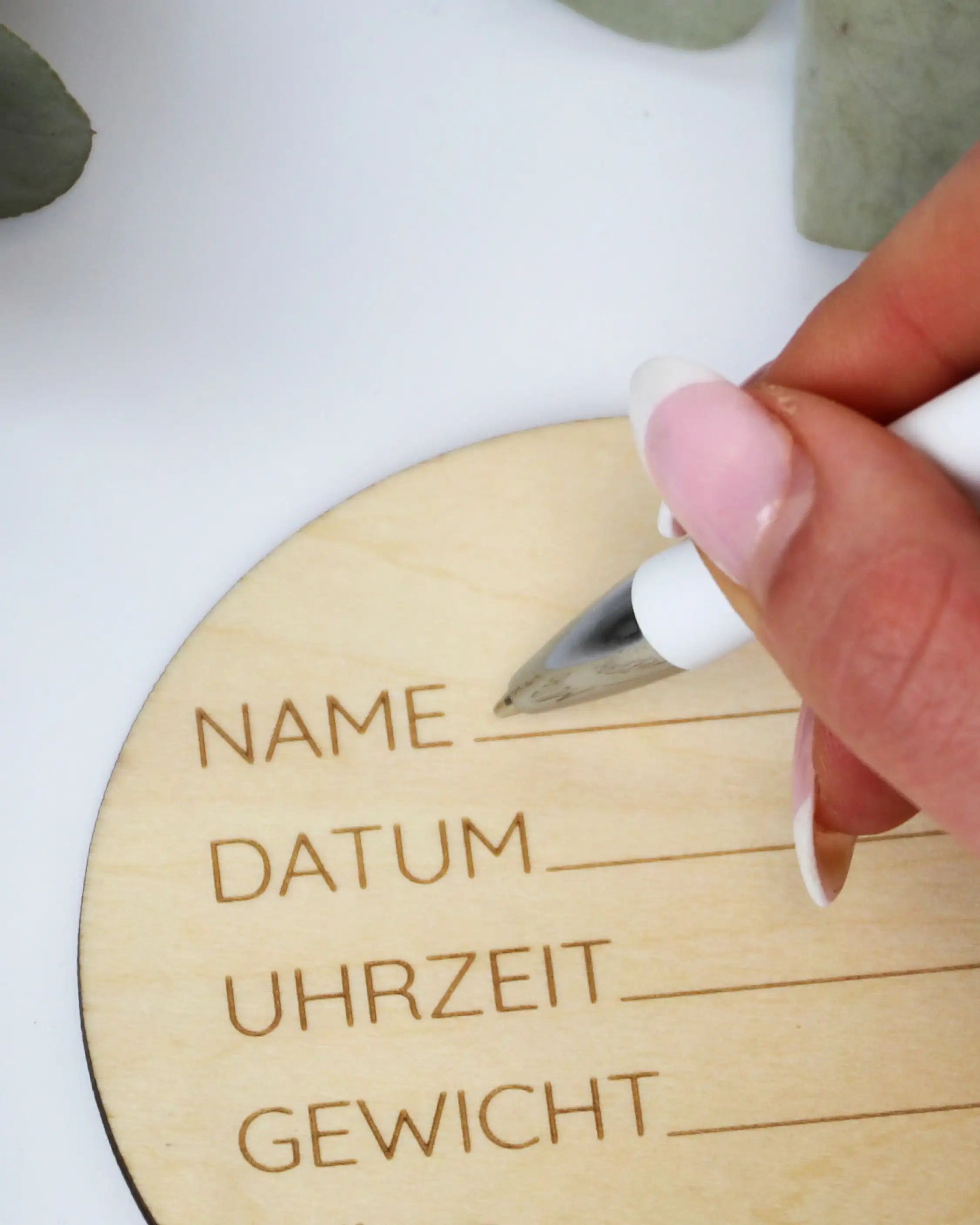 Nahaufnahme eines Holzmeilensteins mit Feldern für Name, Datum, Uhrzeit & Gewicht. Hand trägt Infos ein, Blätter im Hintergrund.