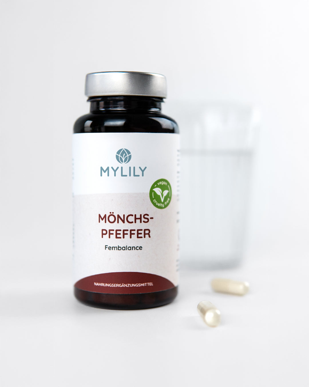 MYLILY Mönchspfeffer „Fembalance" Nahrungsergänzungsmittel in brauner Glasflasche mit veganem und cruelty-free Siegel, neben Wasserglas und zwei Kapseln auf hellem Untergrund