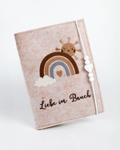 Beige Filz-Mutterpasshülle mit bunter Stickerei, Perlengummi und Herzanhänger, leicht geneigt vor weißem Hintergrund.