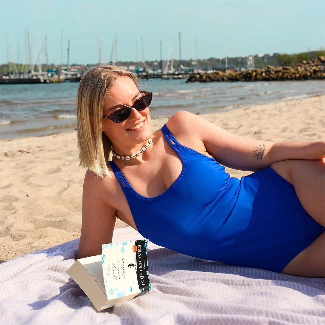 Eine Frau liegt sorglos am Strand und lächelt zufrieden  mit Buch in der Hand. #farbe_royalblau