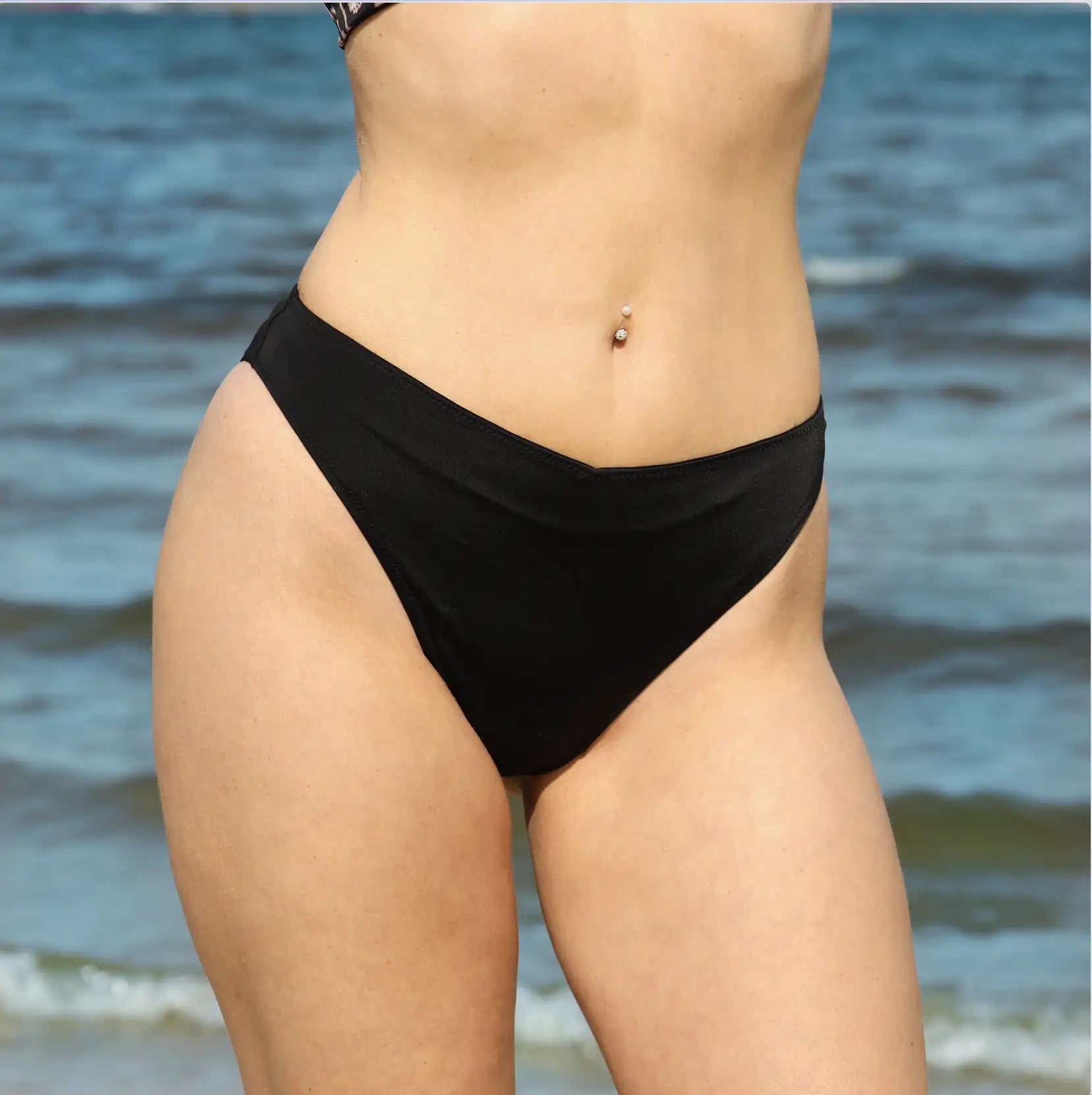 Junge Frau mit Bauchpiercing posiert in einer schwarzen Perioden Bikini Hose am Meer. 